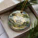 A decorative ornament with a landscape scene featuring a mushroom rock formation, surrounded by pine branches and greenery.