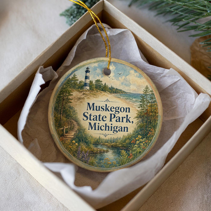A round ornament with a lighthouse and a scenic landscape, placed in a box with a white tissue paper lining.