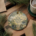 A decorative ornament with a painting of a house and a river, surrounded by greenery, hangs on a wooden surface.
