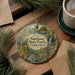 A decorative ornament featuring a scenic forest landscape with a waterfall, surrounded by pine branches and a cup of coffee on a wooden surface.