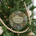 A round ornament hanging on a Christmas tree, featuring a scenic landscape with a bridge, trees, and a bird.