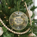 A round ornament hanging on a Christmas tree, featuring a scenic illustration of a park with a bridge and a church.