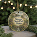 A round ornament with a scenic illustration of a park, featuring a bridge, trees, and a church, is displayed in front of a blurred background with twinkling lights.