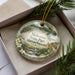 A decorative ornament in a box, featuring a scenic landscape with a building, trees, and water.