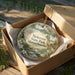 A decorative ornament in a box, featuring a scenic landscape with a castle, trees, and a stream.