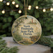 A round ornament with a scenic image of a lighthouse and a bird, surrounded by greenery, is displayed on a surface with Christmas lights in the background.