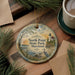 A decorative ornament featuring a lighthouse and a serene landscape, with the text "North Point State Park, Maryland" prominently displayed. The ornament is placed on a wooden surface, surrounded by pine branches and a cup of coffee.