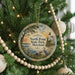 A round ornament hanging on a Christmas tree, featuring a scenic landscape with a lighthouse and a bird in the foreground.