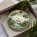 A decorative ornament in a box, featuring a scenic landscape with palm trees, a body of water, and a bird.