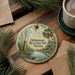 A round ornament with a painting of a river and palm trees, surrounded by greenery, is displayed on a wooden surface alongside a cup of coffee and some papers.
