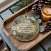 A decorative ornament on a wooden tray, featuring a house and greenery, with a candle and pine cones nearby.