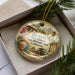A decorative ornament with a vintage-style illustration of a historical building and landscape, placed in a box with pine branches.