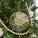 A round ornament hanging on a Christmas tree, featuring a scenic landscape with a cabin and a lake.