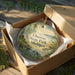 A round ornament in a brown box, featuring a scenic landscape with pine trees, a lake, and mountains, with the text "Onaway State Park, Michigan" printed on it.