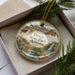 A decorative ornament with a landscape scene featuring a house, mountains, and a waterfall, placed inside a box with pine branches.