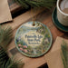 A decorative ornament featuring a scenic landscape of Paintsville Lake State Park, Kentucky, with a bird and flowers, is displayed on a wooden surface alongside a cup of coffee and some papers.