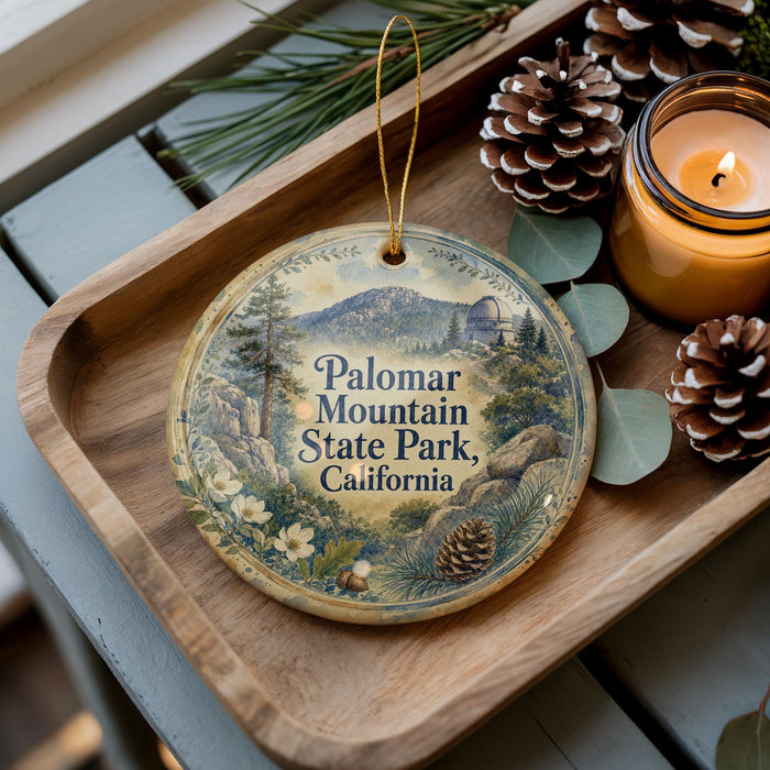 A decorative ornament with a scenic mountain landscape and pine cones sits on a wooden tray, accompanied by a lit candle and pine cones.
