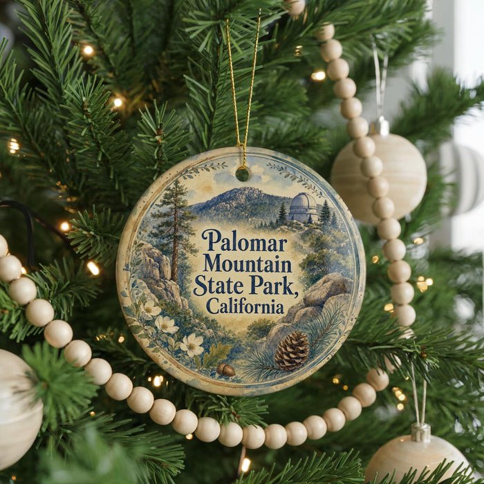 A round ornament hanging on a Christmas tree, featuring a scenic landscape with mountains, pine trees, and a pine cone.
