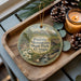 A decorative ornament featuring a scenic landscape of Panola Mountain State Park, Georgia, with pine cones and a lit candle on a wooden tray.