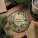 A circular ornament with a scenic landscape of Penwood State Park, Connecticut, featuring pine trees, a river, and a cabin, is displayed on a wooden surface alongside a cup of coffee and some papers.
