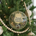 A round ornament on a Christmas tree, featuring a scenic landscape with a winding path, pine trees, and a body of water.