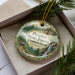 A decorative ornament in a box, featuring a scenic landscape with a waterfall and pine trees.