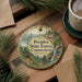 A round ornament with a scenic forest landscape and a waterfall, featuring the text "Peoples State Forest, Connecticut" on it, is displayed on a wooden surface alongside a cup of coffee and some pine branches.