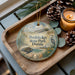 A decorative ornament with a beach scene and a bird, placed on a wooden tray next to a lit candle and pine cones.