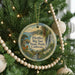 A round ornament hanging on a Christmas tree, featuring a scenic landscape with a river, mountains, and trees.