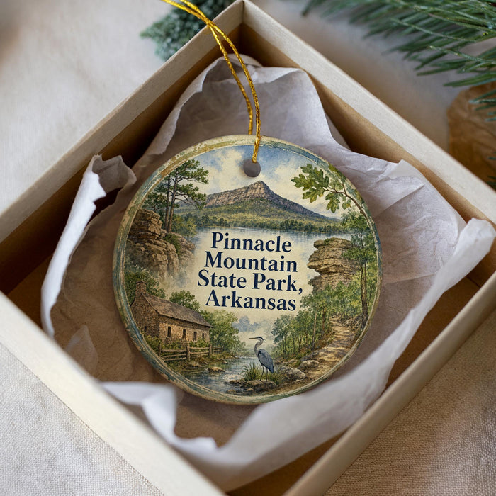 A decorative ornament in a box, featuring a scenic landscape with a mountain, cabin, and bird, along with the text "Pinnacle Mountain State Park, Arkansas".