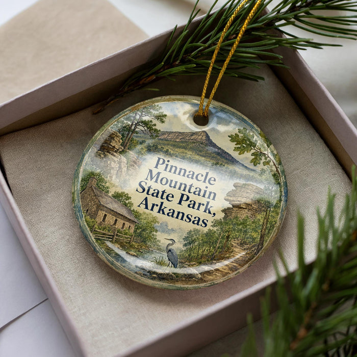 A decorative ornament in a box, featuring a scenic landscape with mountains, trees, and a bird, along with the text "Pinnacle Mountain State Park, Arkansas".