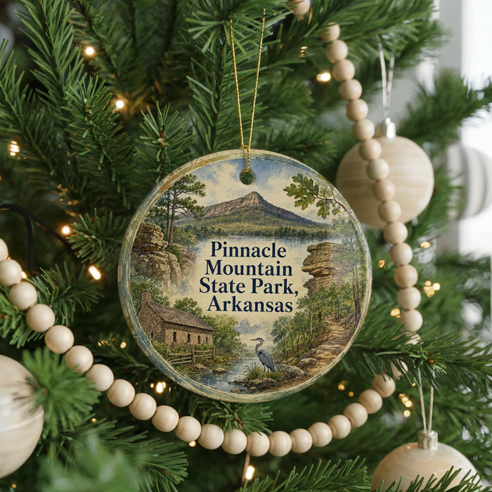 A round ornament hanging on a Christmas tree, featuring a scenic landscape with mountains, trees, and a bird.