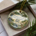 A decorative ornament in a box, featuring a scenic landscape with a river, trees, and wildlife, including a deer and a bird.