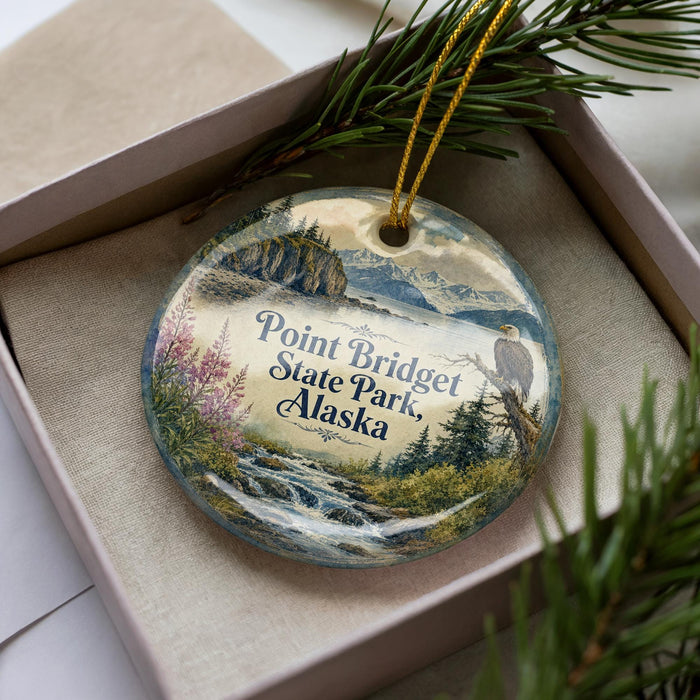 A decorative ornament in a box, featuring a scenic landscape with mountains, water, and pine trees.