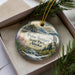 A decorative ornament in a box, featuring a scenic landscape with mountains, water, and pine trees.