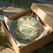 A round ornament in a box, featuring a lighthouse and a scenic landscape, with the text "Point Lookout State Park, Maryland" printed on it.