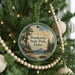 A round ornament hanging on a Christmas tree, featuring a scenic landscape with a cabin and pine trees.