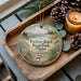 A decorative ornament with a beach scene and a bird is displayed on a wooden tray, accompanied by pine cones and a lit candle.