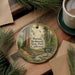 A decorative ornament featuring a scenic image of Portola Redwoods State Park in California, with a path leading through the forest and a bridge in the background. The ornament is placed on a wooden surface, surrounded by pine branches and a cup of coffee.