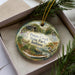 A decorative ornament in a box, featuring illustrations of a deer, a bird, and a bridge, with the text "Potato Creek State Park, Indiana" printed on it.