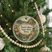 A round ornament hanging on a Christmas tree, featuring a scenic landscape with a river, bridge, and house.