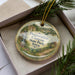 A decorative ornament in a box, featuring a scenic landscape with a waterfall and greenery, with the text "Preparation Canyon State Park, Iowa" printed on it.