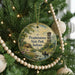 A round ornament hanging on a Christmas tree, featuring a scenic landscape with a river, a lighthouse, and a small boat.