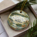 A decorative ornament with a scenic lake and forest landscape, placed in a box with pine branches.