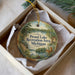 A round ornament with a scenic landscape and text, placed in a box with a white tissue paper lining.