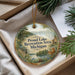 A round ornament with a scenic landscape and text, placed in a box with pine branches.