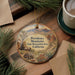 A decorative ornament with a scenic mountain landscape and text, placed on a wooden surface alongside a cup of coffee and some pine branches.