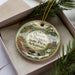 A decorative ornament in a box, featuring a scenic landscape with a waterfall and flowers, with the text "Queen Wilhelmina State Park, Arkansas" printed on it.