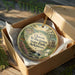 A decorative ornament in a box, featuring a scenic landscape with a river, trees, and flowers.