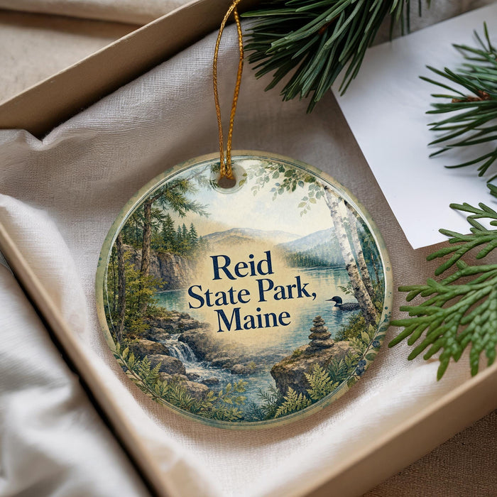 A decorative ornament with a scenic landscape of Reid State Park, Maine, featuring a waterfall, pine trees, and mountains.